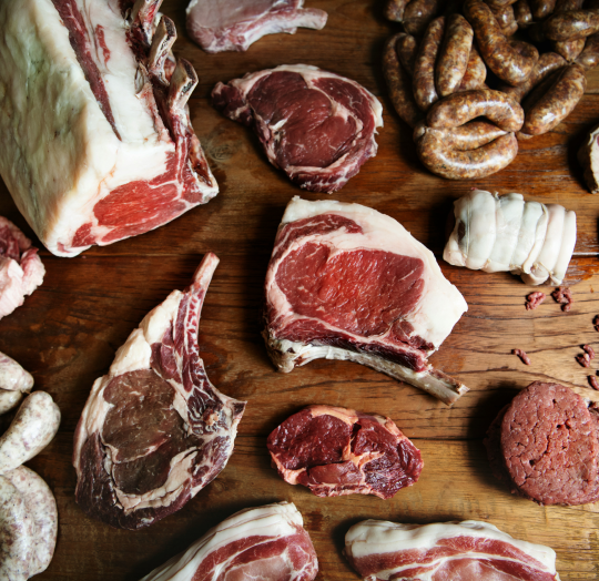 A top down view of assorted fresh raw meat cuts arranged on a wooden table including thick ribeye steaks sausages and a bone in roast representing the Butcher and Creamery collection.