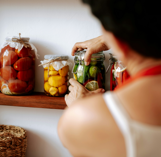 A person placing a jar of homemade pickles onto a wooden shelf next to preserved tomatoes and yellow squash representing the local goods in the Farm Pantry collection.
