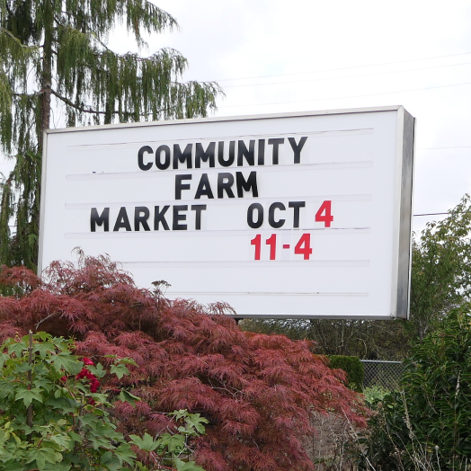 A white community farm market sign announcing an October event in the Fraser Valley, inviting neighbours to shop for local Farmbox produce and artisan goods.