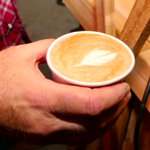 A close up of a hand holding a freshly poured latte with leaf art in a paper cup at the community market and bistro.